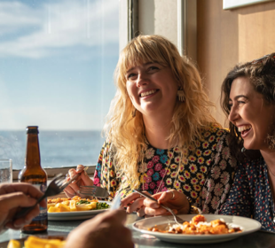 Two women laughing and eating food at a window seat looking out to sea