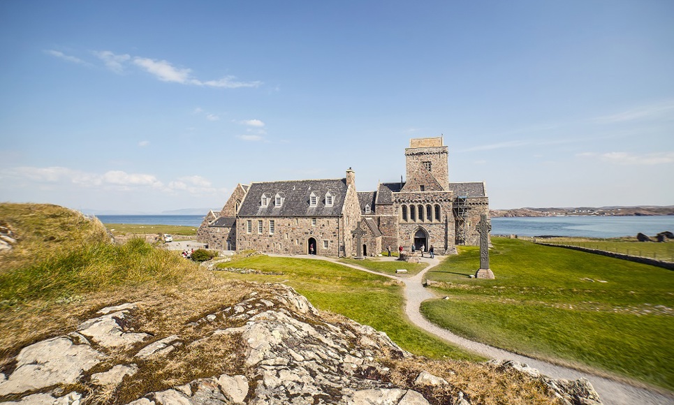 The ancient stone building of Iona Abbey taking from a rocky hilltop with the costline and water in the background.