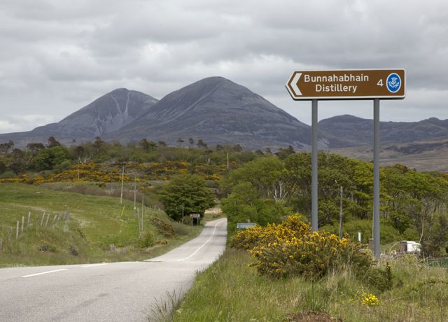 The Paps of Jura hills in the background with an quiet road and sign for an Islay whiskey distillery in the foreground