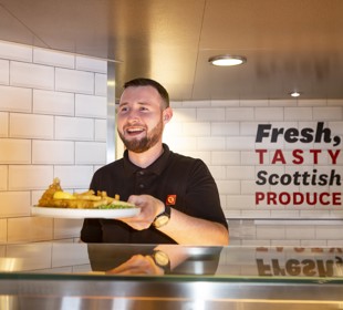 a server passing a plate of fish, fries and peas to a customer from the servery