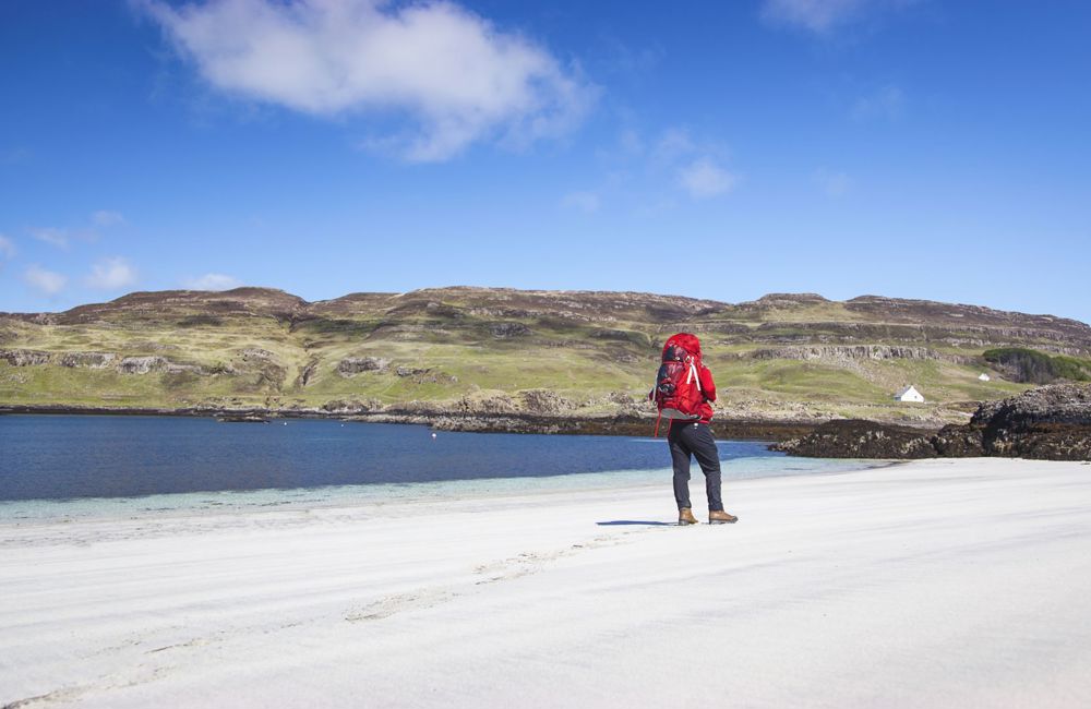 A hiker with a vibrant red jacket, standing on a white sand beach under a blue sky with gentle green hills in the background.