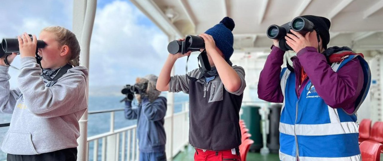 Group of kids outside on a ferry looking out to sea with binoculars 