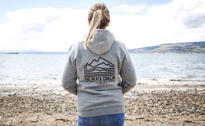 A woman on a beach, wearing a grey CalMac hoodie with the writing The Black Cuillin. She is staring to the sea in front of her.