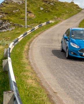 A blue car driving down a single track, coastal road