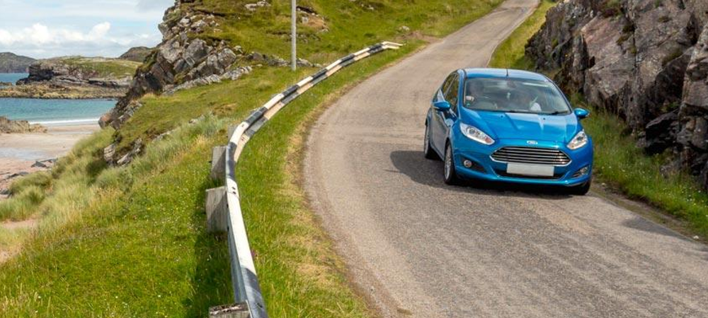 A blue car driving down a single track, coastal road