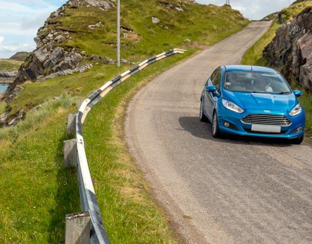 A blue car driving down a single track, coastal road