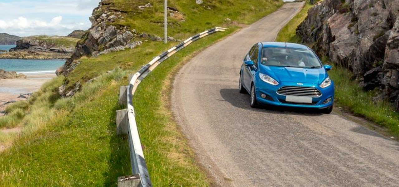 A blue car driving down a single track, coastal road