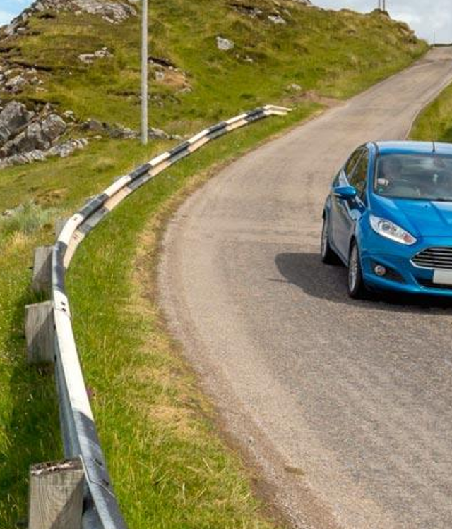 A blue car driving down a single track, coastal road