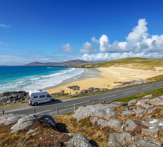 A view from a rock hill across a road to a golden beach with vibrant blue sea and sky