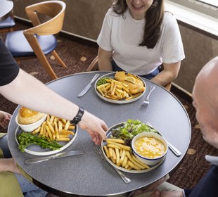 someone placing macaroni cheese, fries and salad in front of a man who's sitting at a table with two females