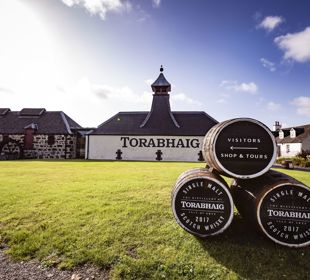 Outside view of Torabhaig Distillery with a white front and dark chimney, with 3 whisky barrels on the grass in the foreground.