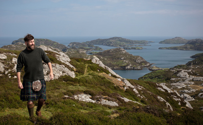 The Hebridean Baker wearing a kilt on top of a mountain with view out to sea behind him