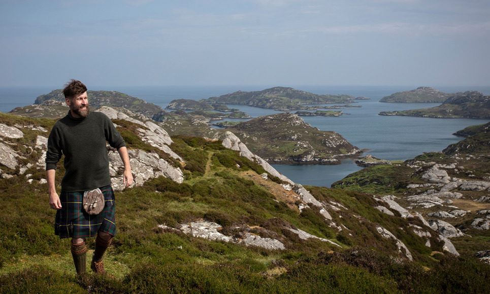 The Hebridean Baker wearing a kilt on top of a mountain with view out to sea behind him