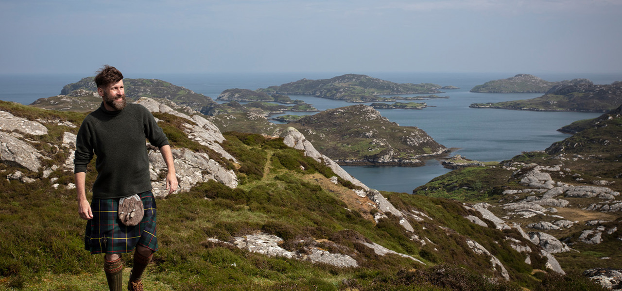 The Hebridean Baker wearing a kilt on top of a mountain with view out to sea behind him